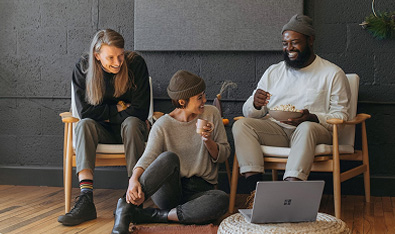 Trois personnes en discussion détendue dans un espace moderne : une personne assise sur une chaise, une autre assise par terre, et une troisième assise sur un fauteuil avec un ordinateur portable ouvert devant eux, dans une ambiance chaleureuse et conviviale.